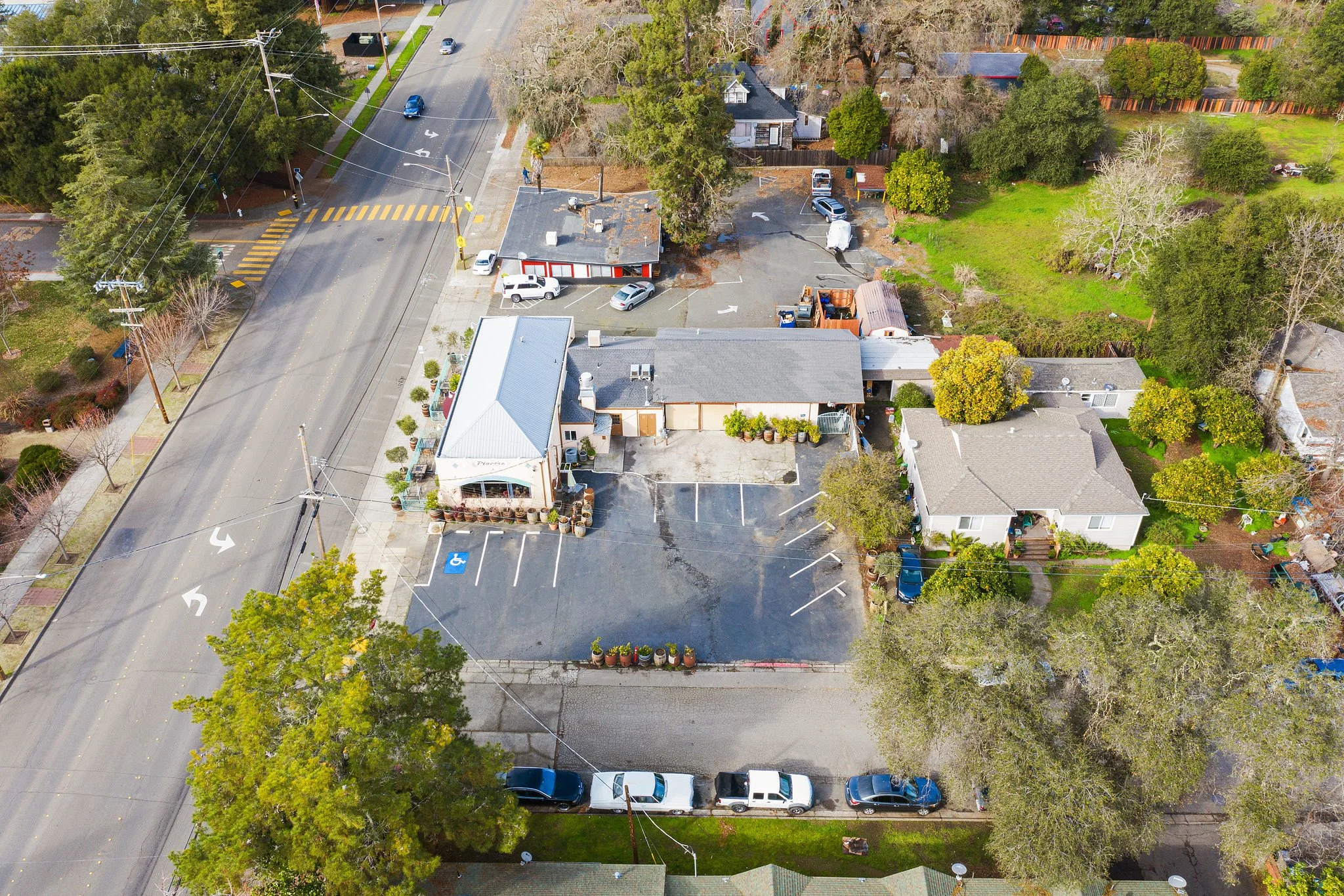 Aerial view of a small commercial building with outdoor seating and parking lot, adjacent to residential houses and green spaces, with a busy street and power lines.