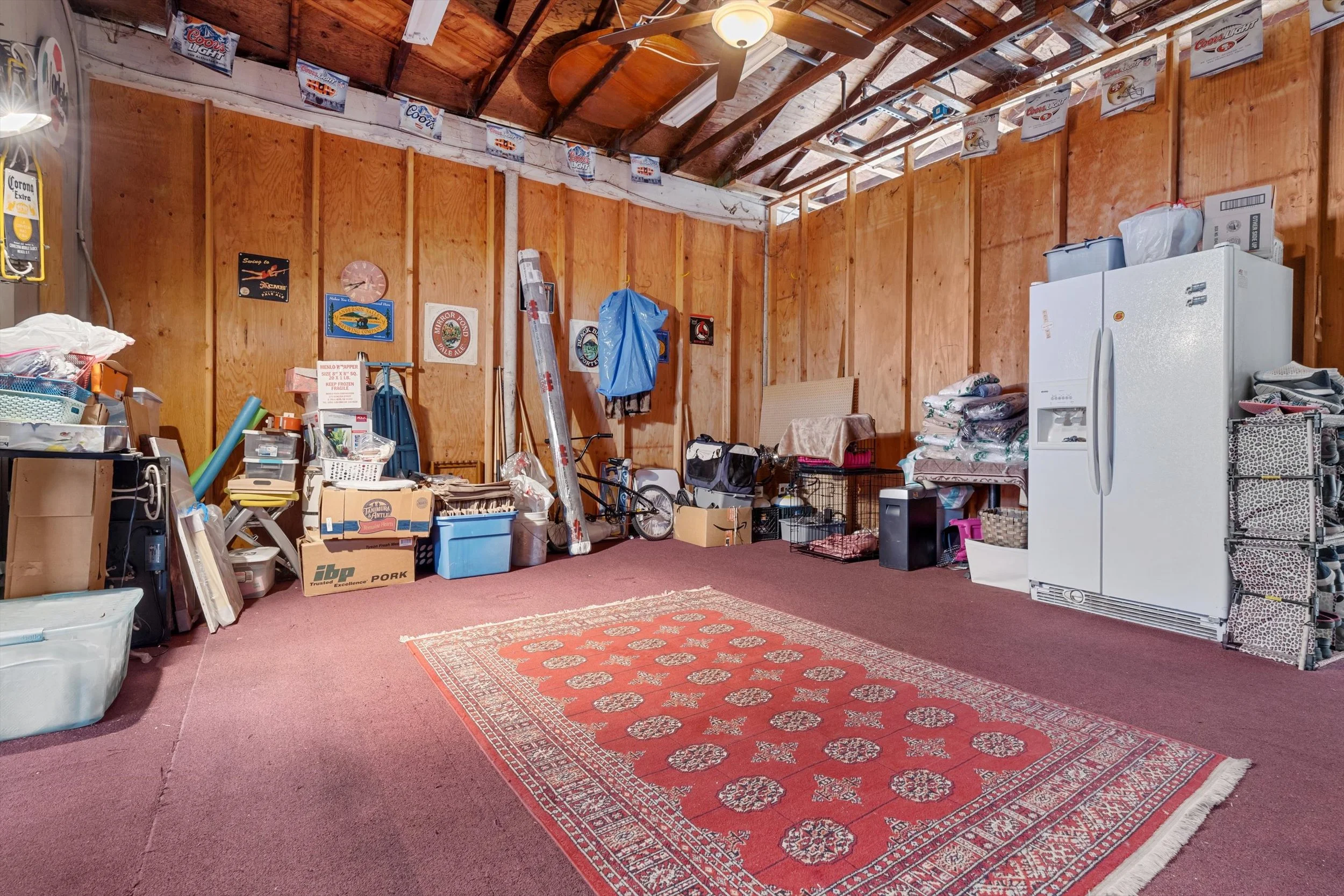 A garage with unfinished wood-paneled walls and roof, filled with various items including boxes, tools, a bicycle, and a refrigerator, on a red carpeted floor with a central patterned rug.