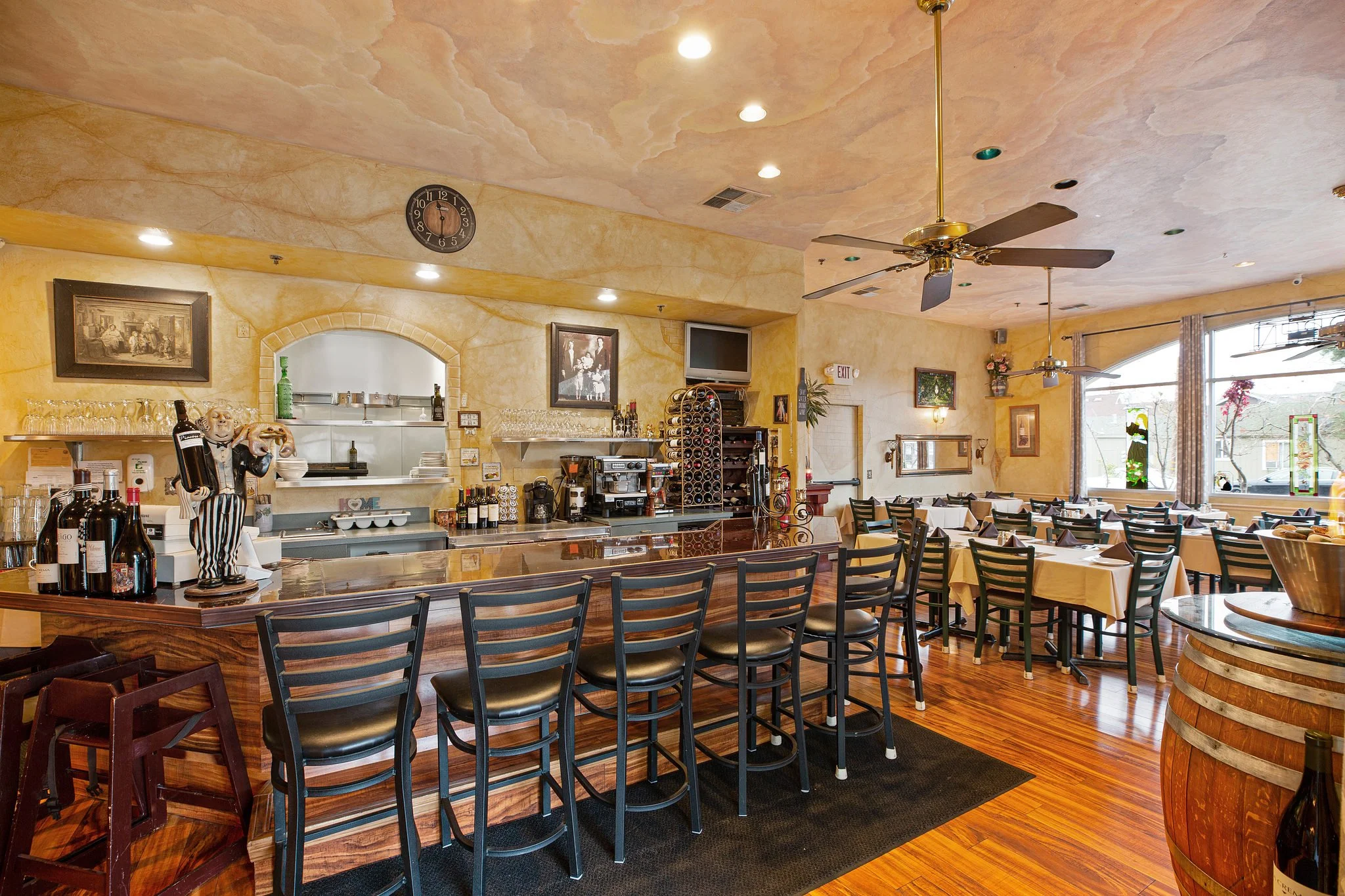 Interior of a cozy restaurant with a bar area, wine bottles, and wooden furniture, including chairs and tables set for dining near large windows with natural light.