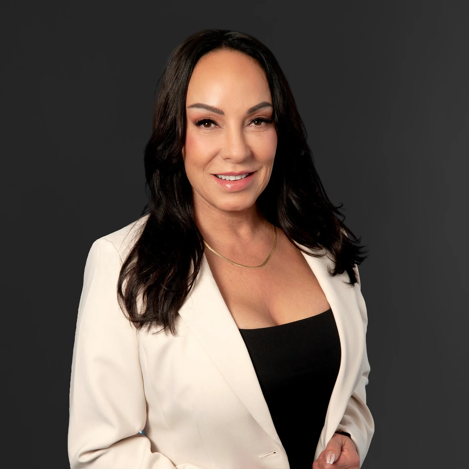 Professional woman with dark hair wearing a white blazer over a black top, smiling against a dark background.