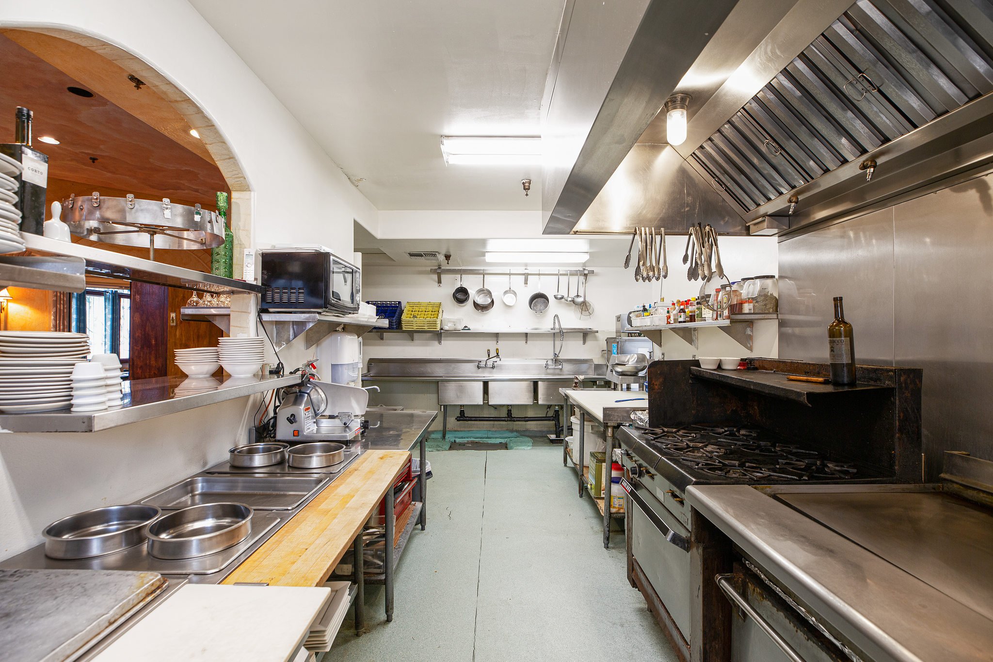 Commercial kitchen with stainless steel appliances, cooking utensils hanging above a stove, shelves with dishes, and a work countertop.