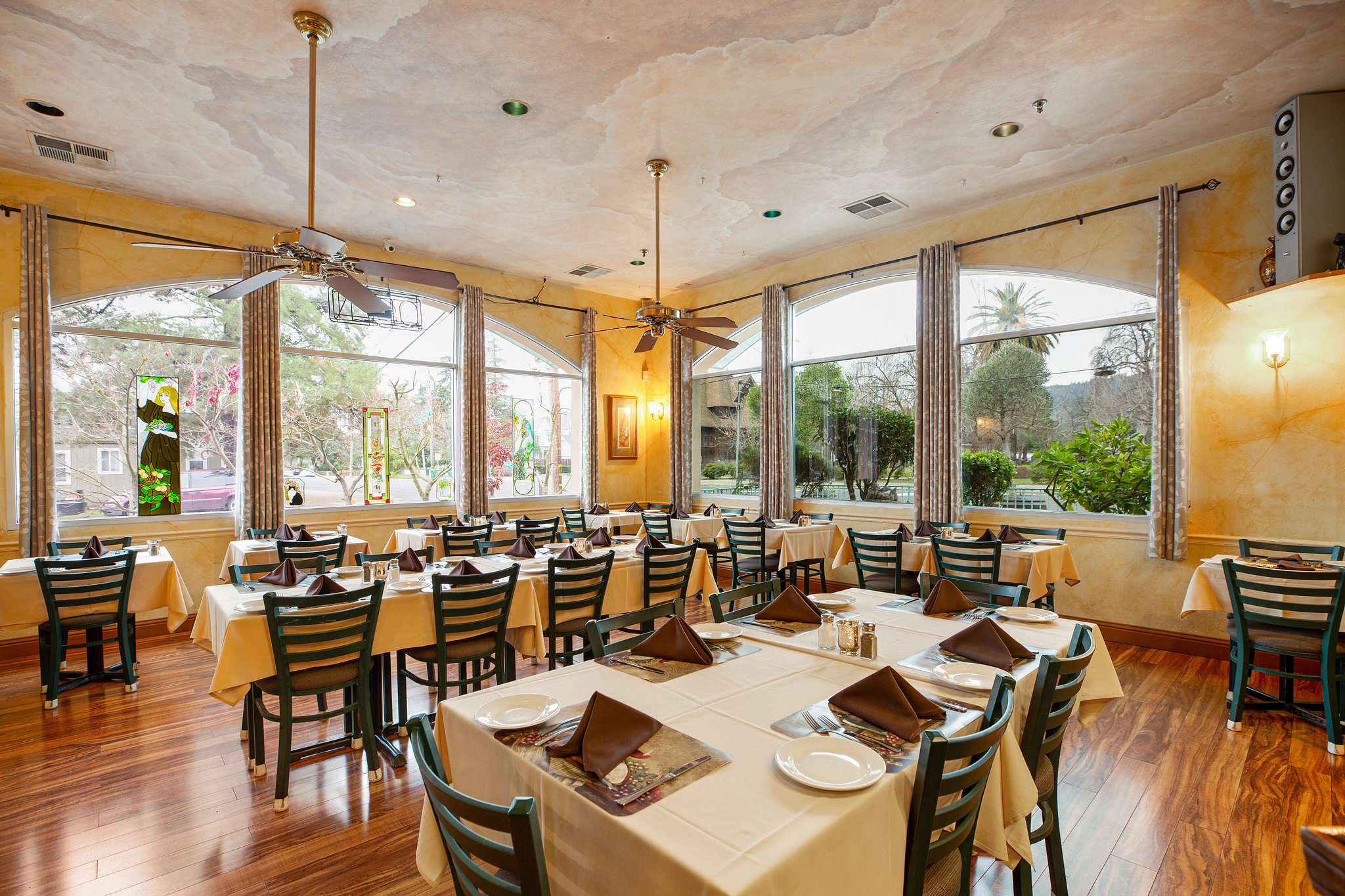 Restaurant with large windows, set dining tables with beige tablecloths, brown napkins, and place settings, wooden floors, ceiling fans, and decorative stained glass panels on the windows.