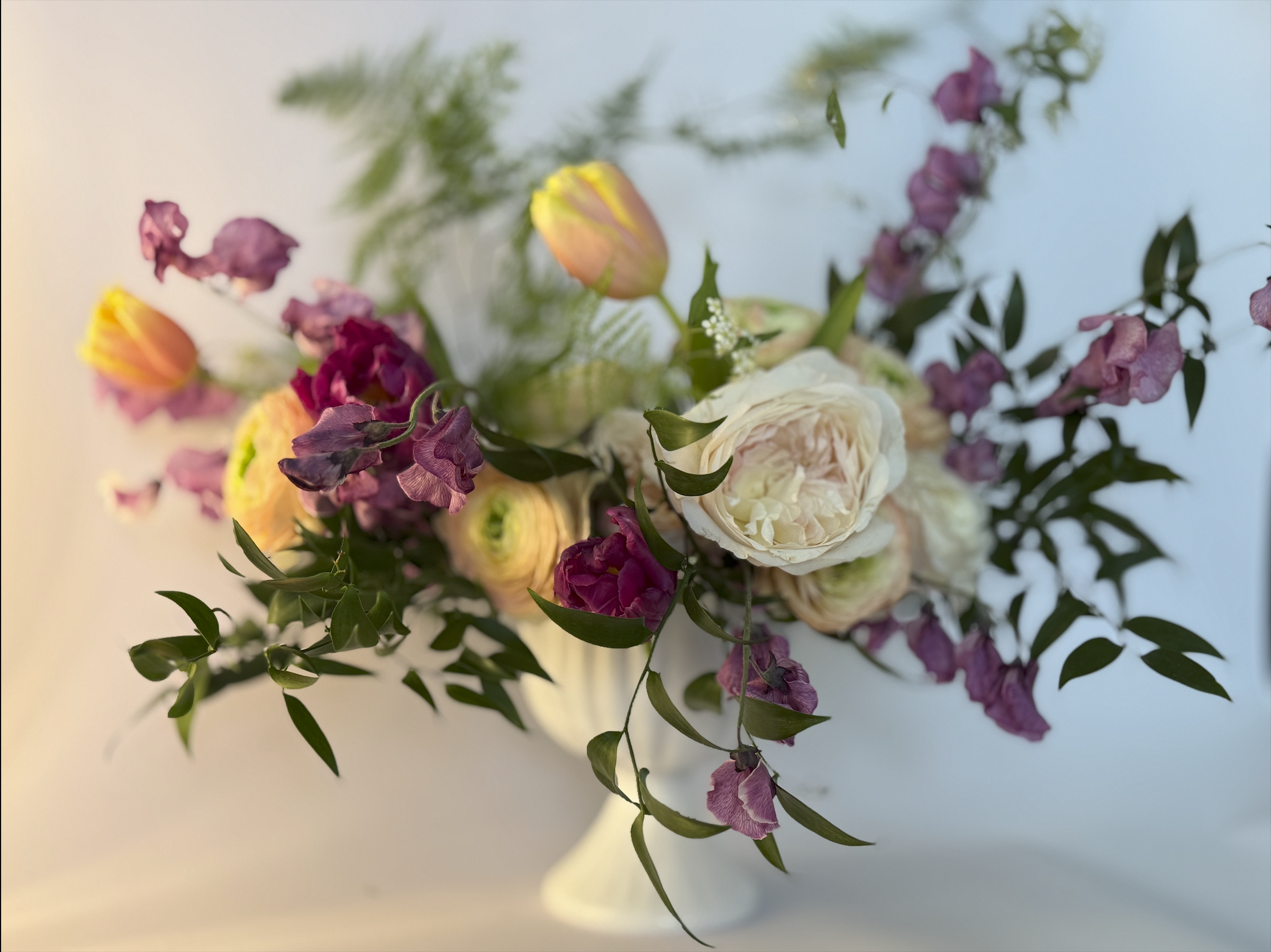 A bouquet of mixed flowers including white and pink peonies, purple snapdragons, and yellow tulips with green leaves in a white vase against a soft, light background.