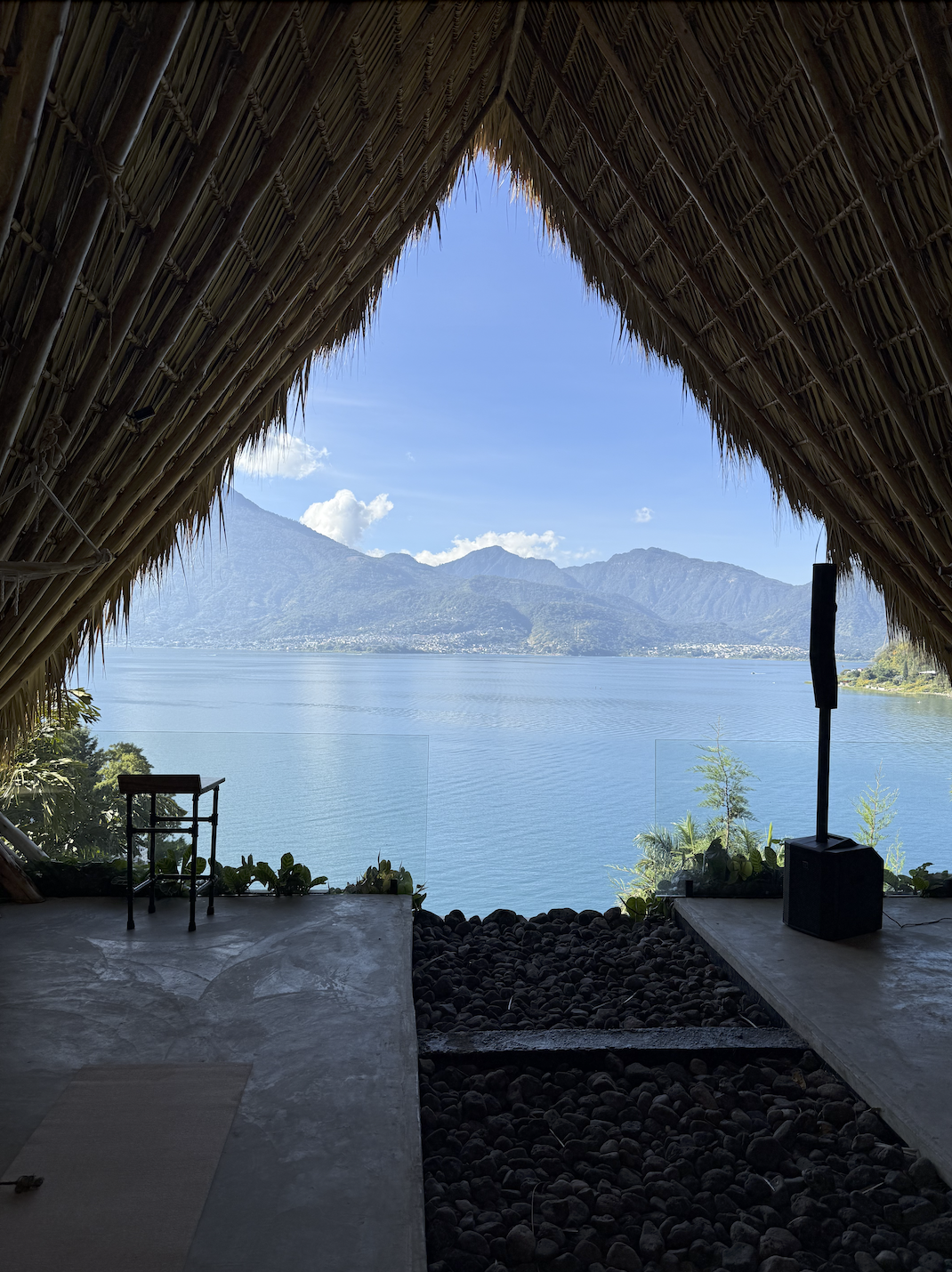 View of a lake and mountains through the opening of a thatched-roof structure with rocks and plants at the edge.