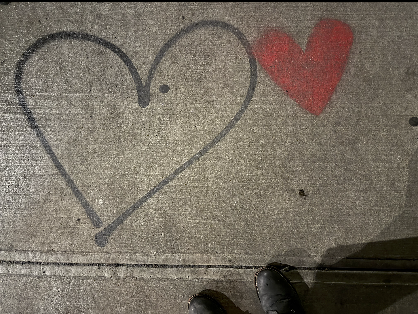 A sidewalk with a chalk drawing of a heart with a smaller heart inside it, and a separate red-painted heart nearby. The photo is taken from above, showing Lisa's shoes. Lisa always takes pictures of hearts out and about when she sees them.