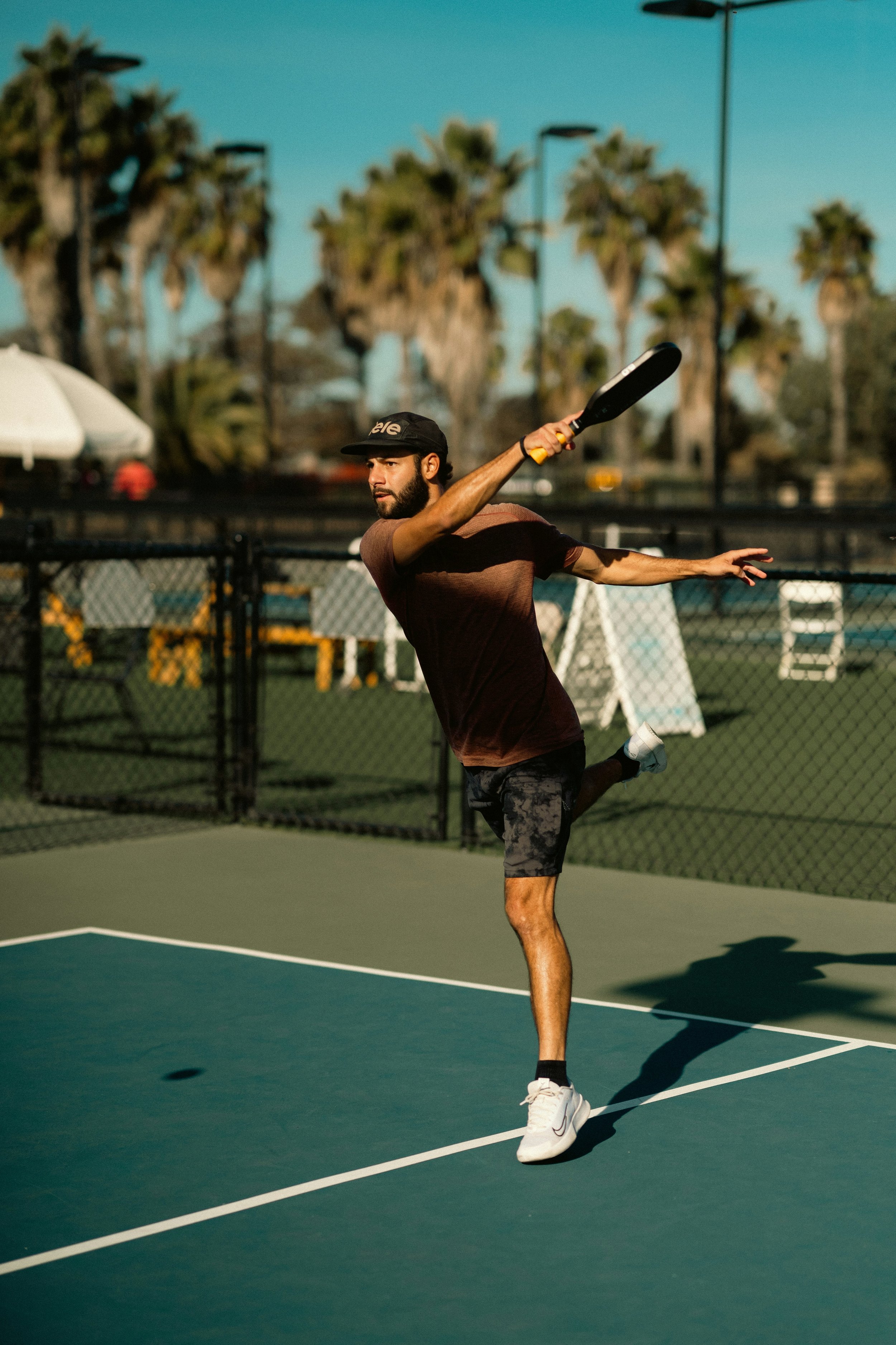 A man playing pickleball on a court, swinging a paddle at a ball. He is wearing a black cap, brown t-shirt, dark shorts, and white sneakers. Palm trees are visible in the background.