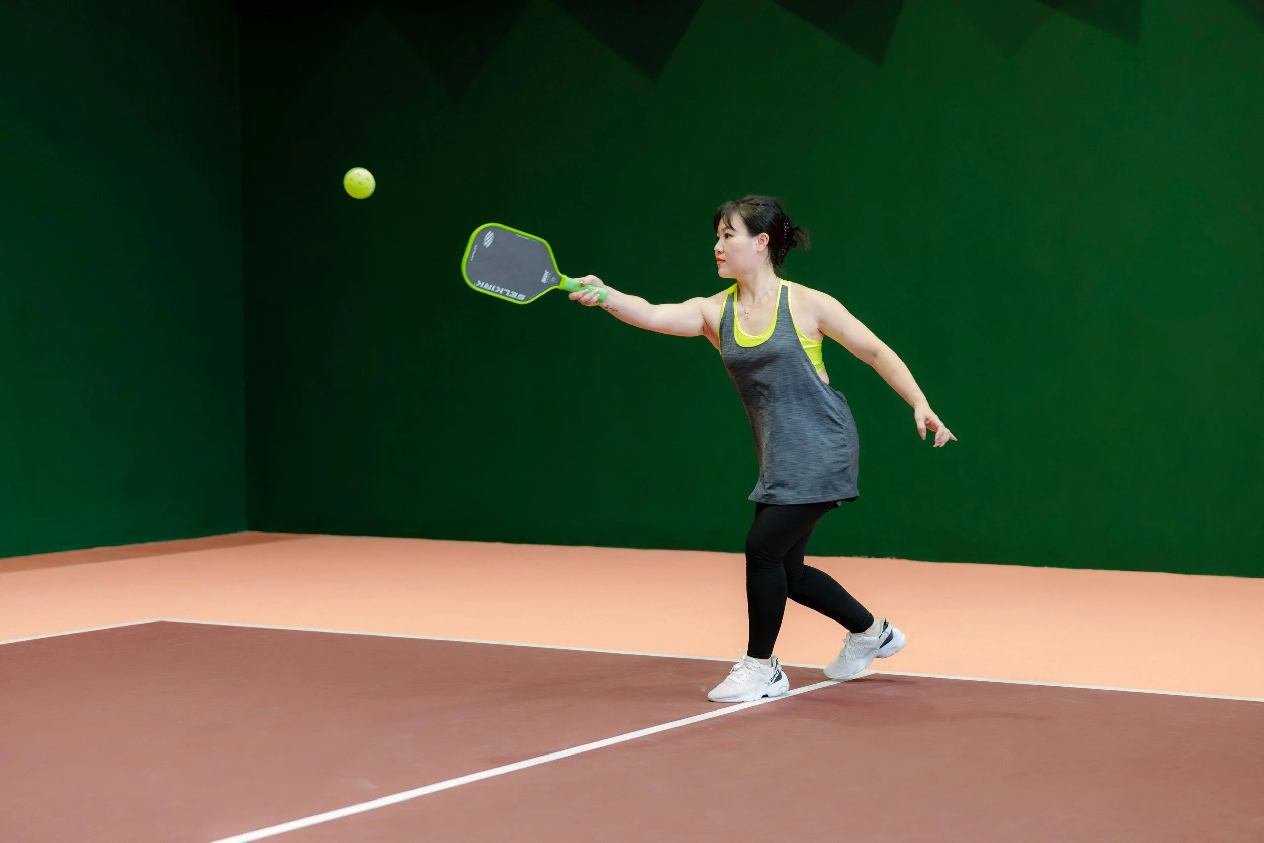 A woman playing pickleball on an indoor court with green walls, hitting a yellow pickleball with a Covert Pickleball paddle.