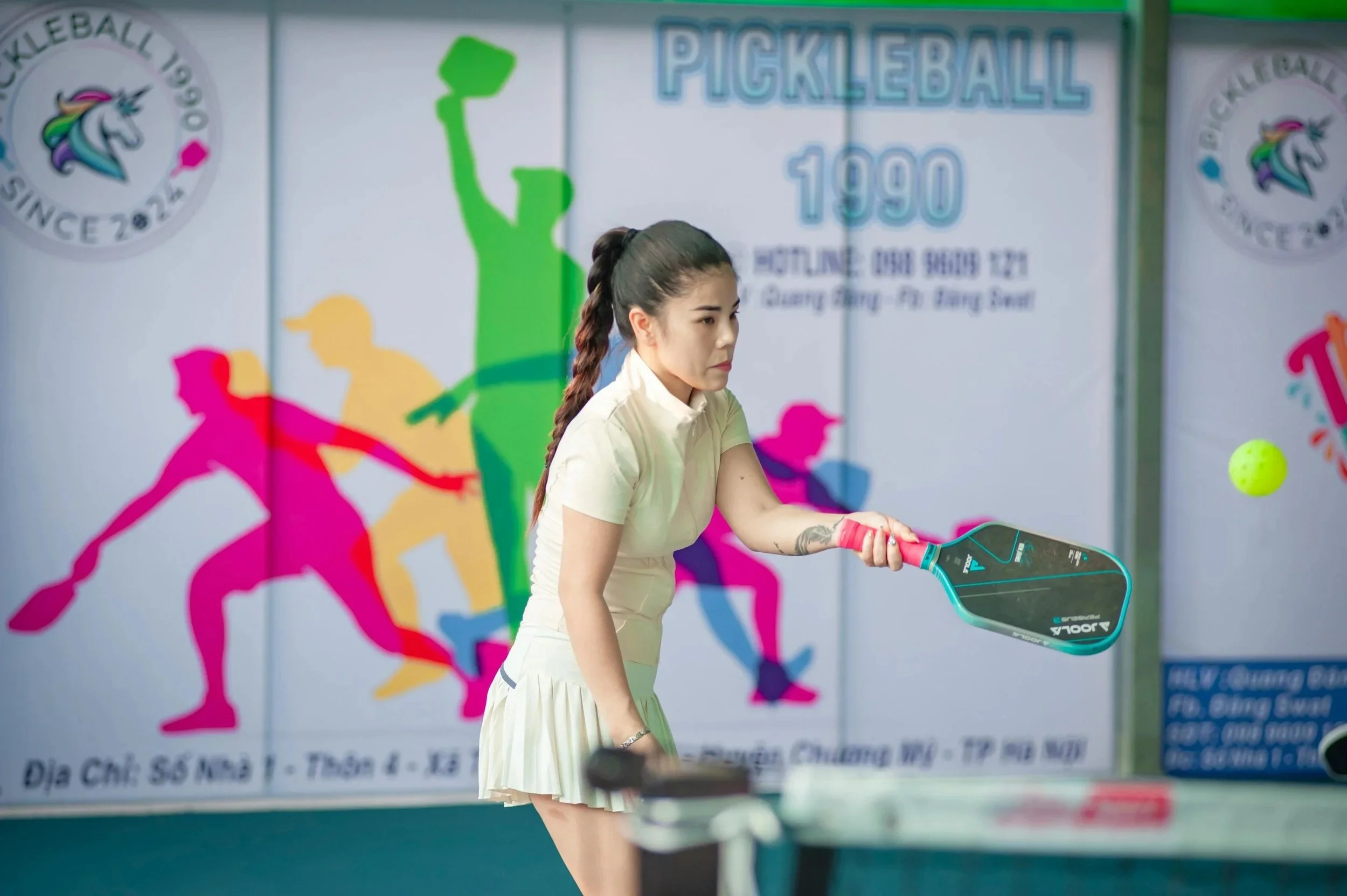 A woman playing pickleball indoors, holding a paddle and looking at a yellow pickleball. The background has a colorful poster with the word "PICKLEBALL".