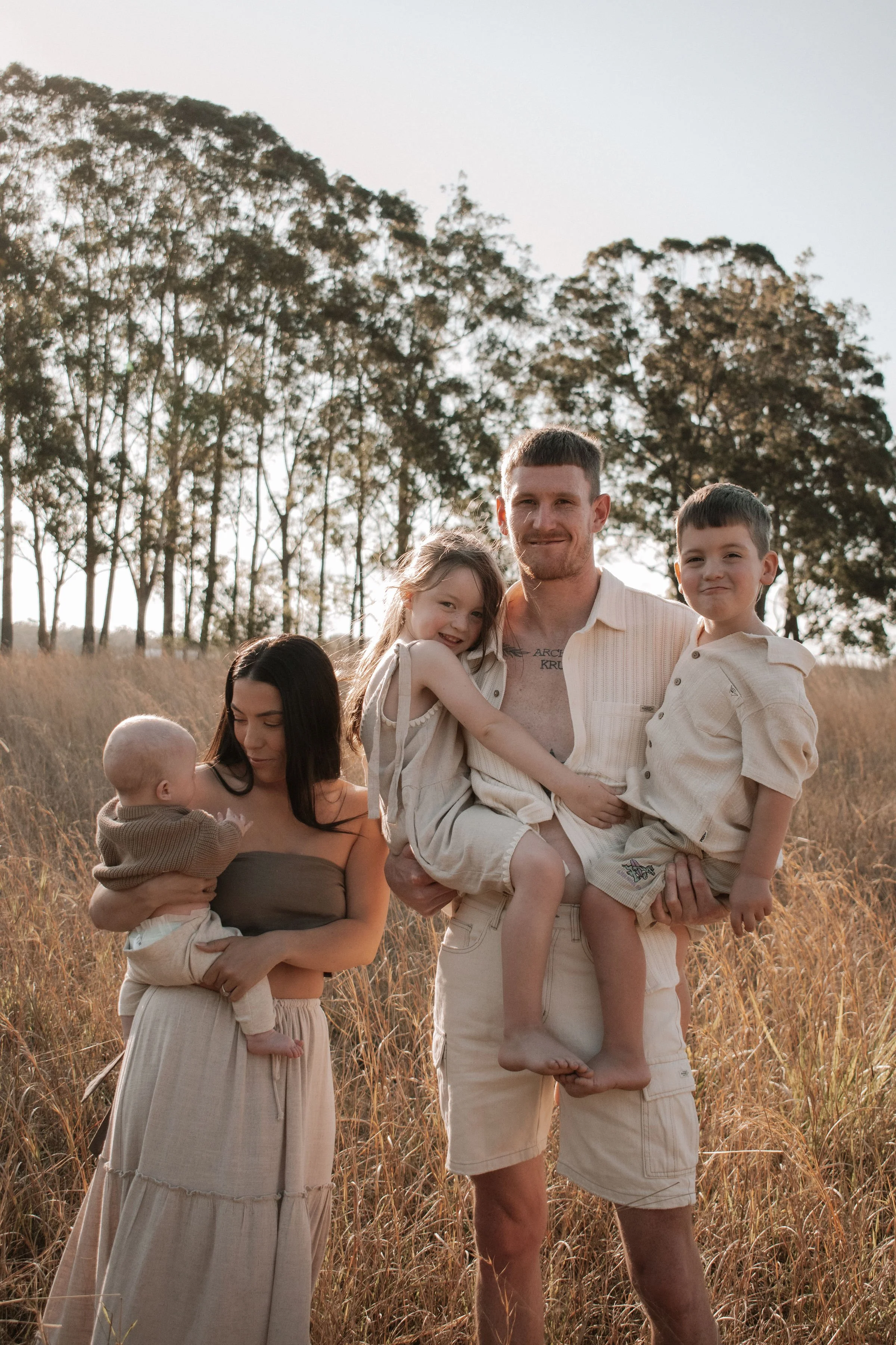 A family of five outdoors in a grassy field with trees in the background, during sunset. The father is holding two children, a girl and a boy, while the mother holds a baby.