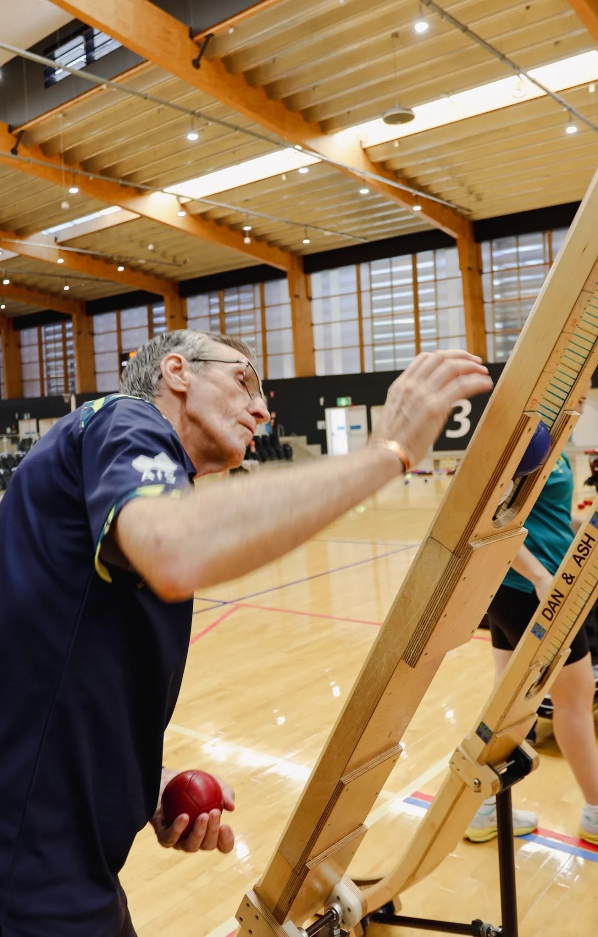 An elderly man in a sports uniform on an indoor court, holding a red boccia ball in his left hand and placing it on a boccia ramp, under a wooden ceiling with lighting.
