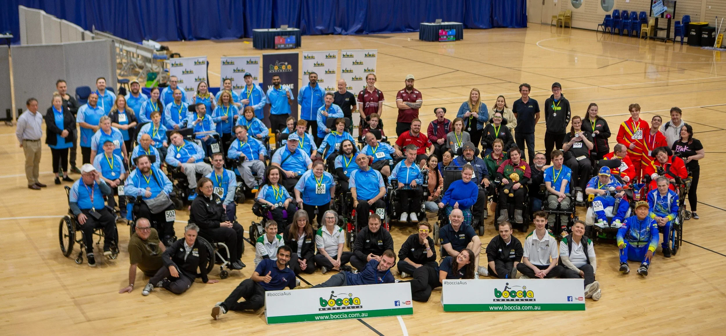 Group photo of athletes, coaches, and staff at a boccia tournament in a gymnasium, with individuals in wheelchair and standing, holding banners with the Boccia Australia logo.
