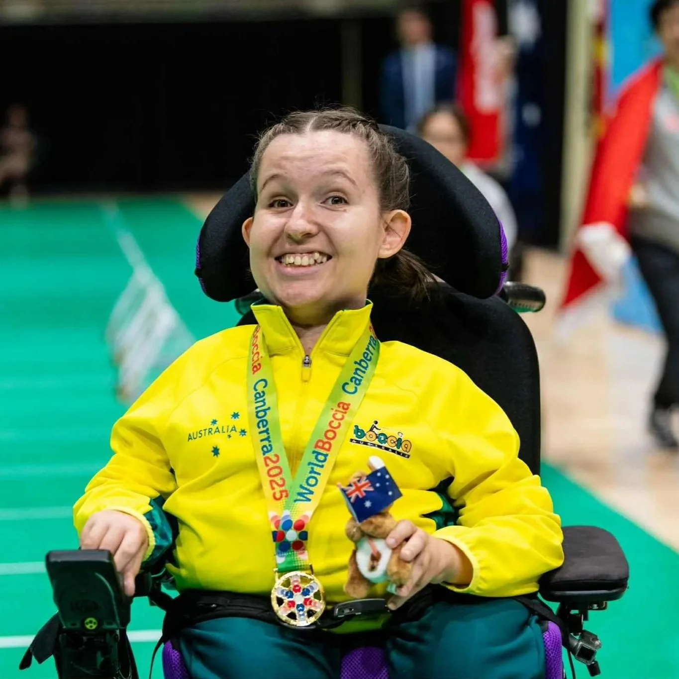 A smiling woman in a yellow jacket sitting in a wheelchair, wearing a medal with 'World Boccia 2025' on it around her neck, holding a small plush toy with an Australian flag. The woman is at an indoor sports event, with a blurred background of people and flags.