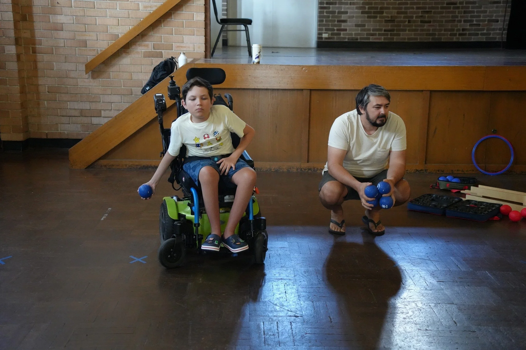 A young boy in a wheelchair holding blue boccia balls next to an adult man crouching with blue boccia balls, in a room with a wooden stage and various boccia equipment on the floor.