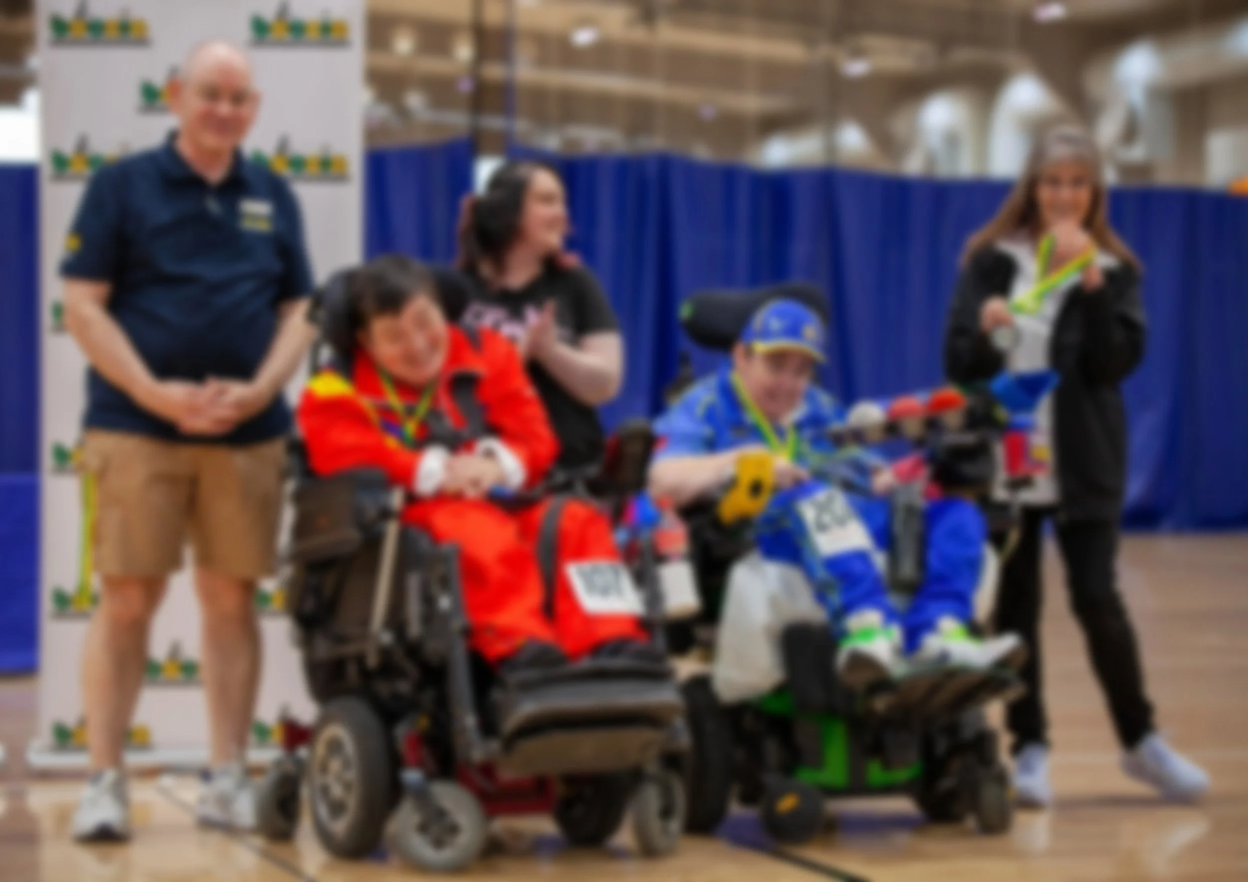 Two children in wheelchairs wearing sports gear, surrounded by smiling adults at an indoor sports event with blue curtains in the background.