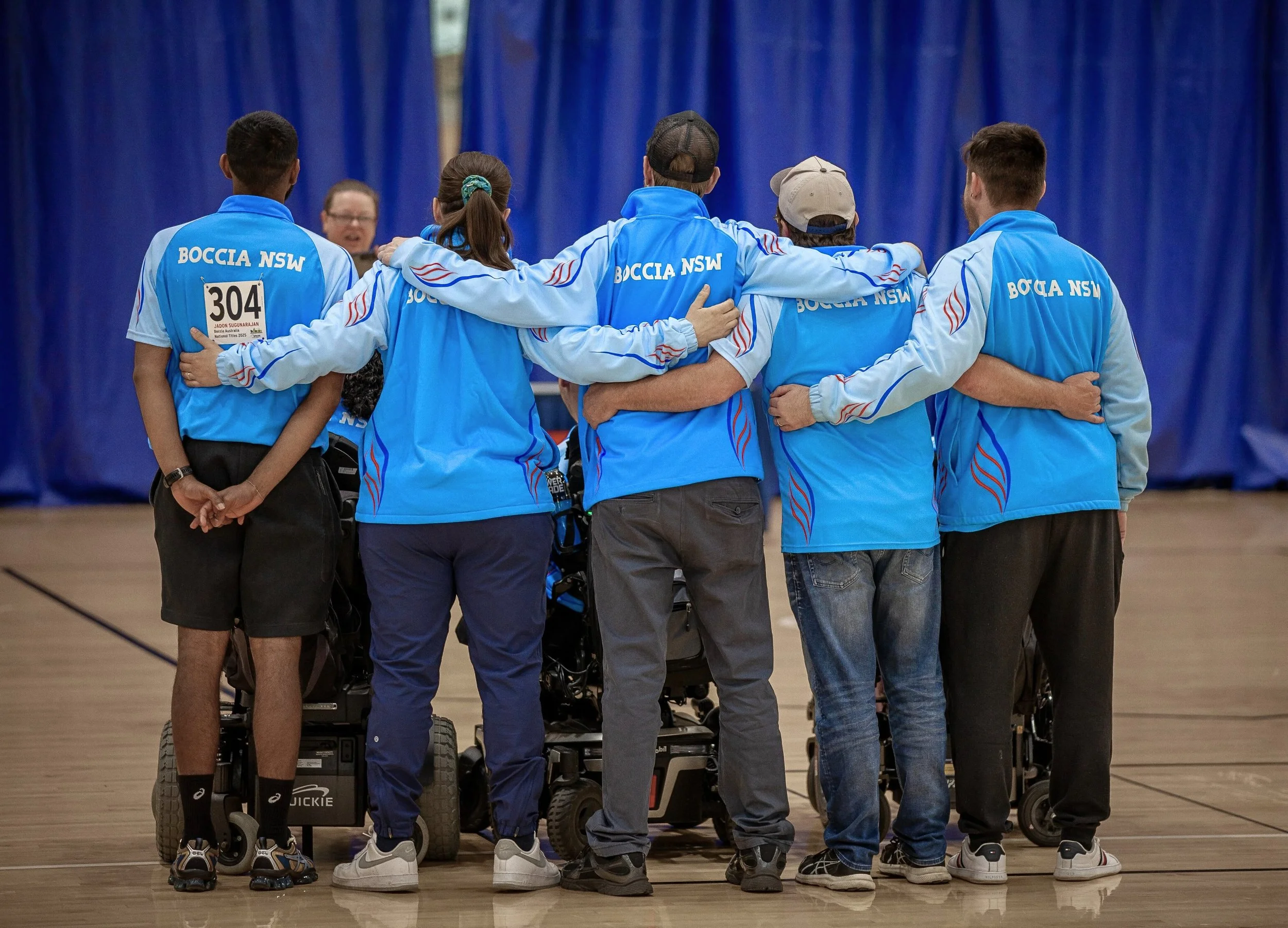 Group of Boccia players in blue uniforms with their arms around each other, standing in a circle on a court, with blue curtains in the background.