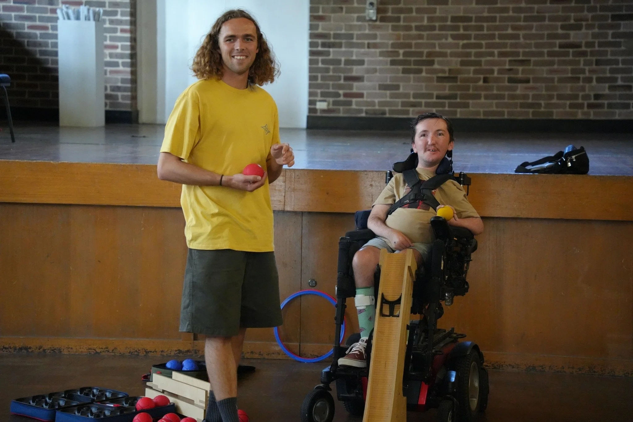 Two young men, one standing and one in a power wheelchair, appear to be playing  game inside a room with brick walls. The man standing is holding a red ball and smiling, while the man in the wheelchair has a yellow ball in his lap. There are various balls and equipment on the floor.