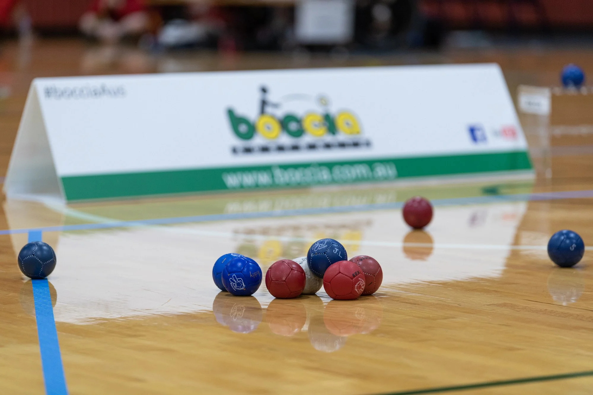Boccia game with red, blue, and white balls on a wooden court with a white barrier in the background.