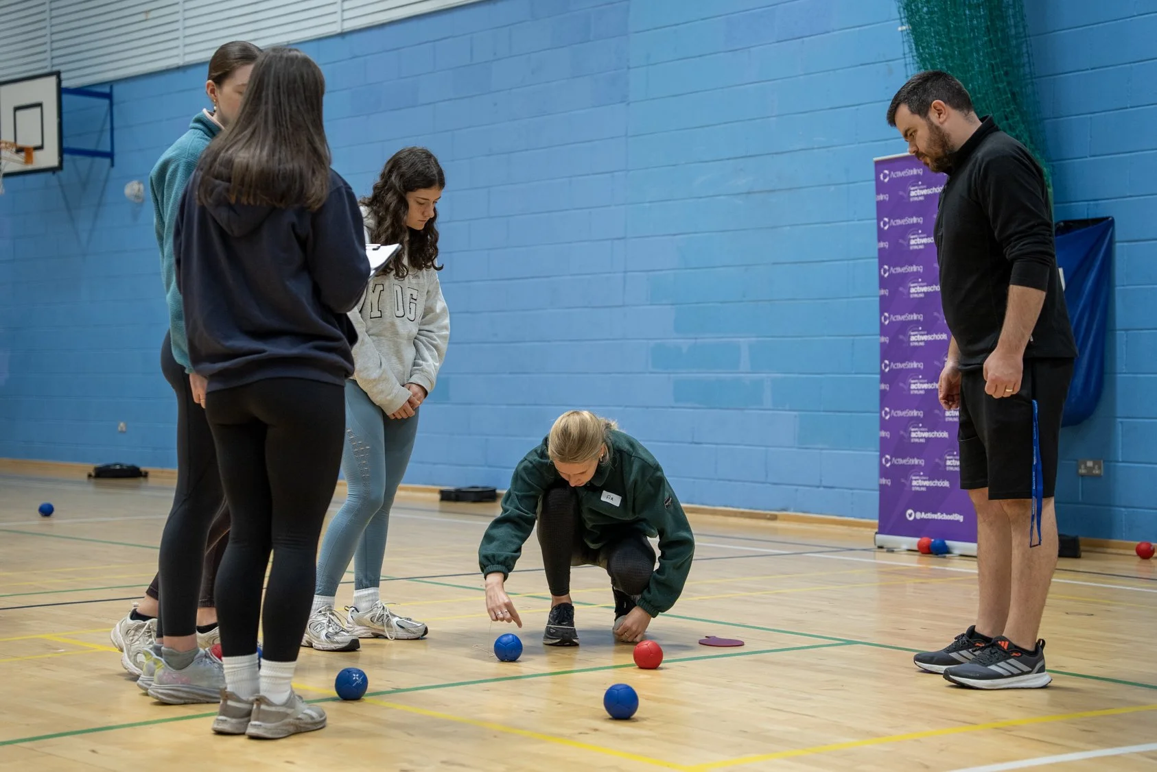 A group of people measuring boccia balls in a gymnasium with a blue wall, with various boccia balls on the floor.