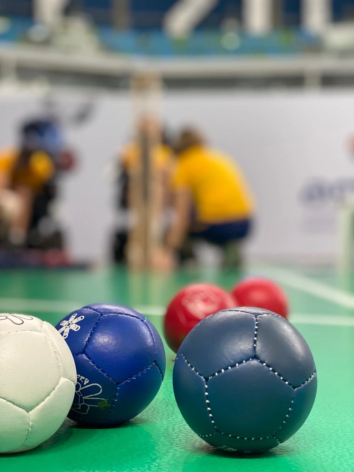 Close-up of colorful boccia balls, set on a green taraflex court with blurred players in yellow and black uniforms in the background.