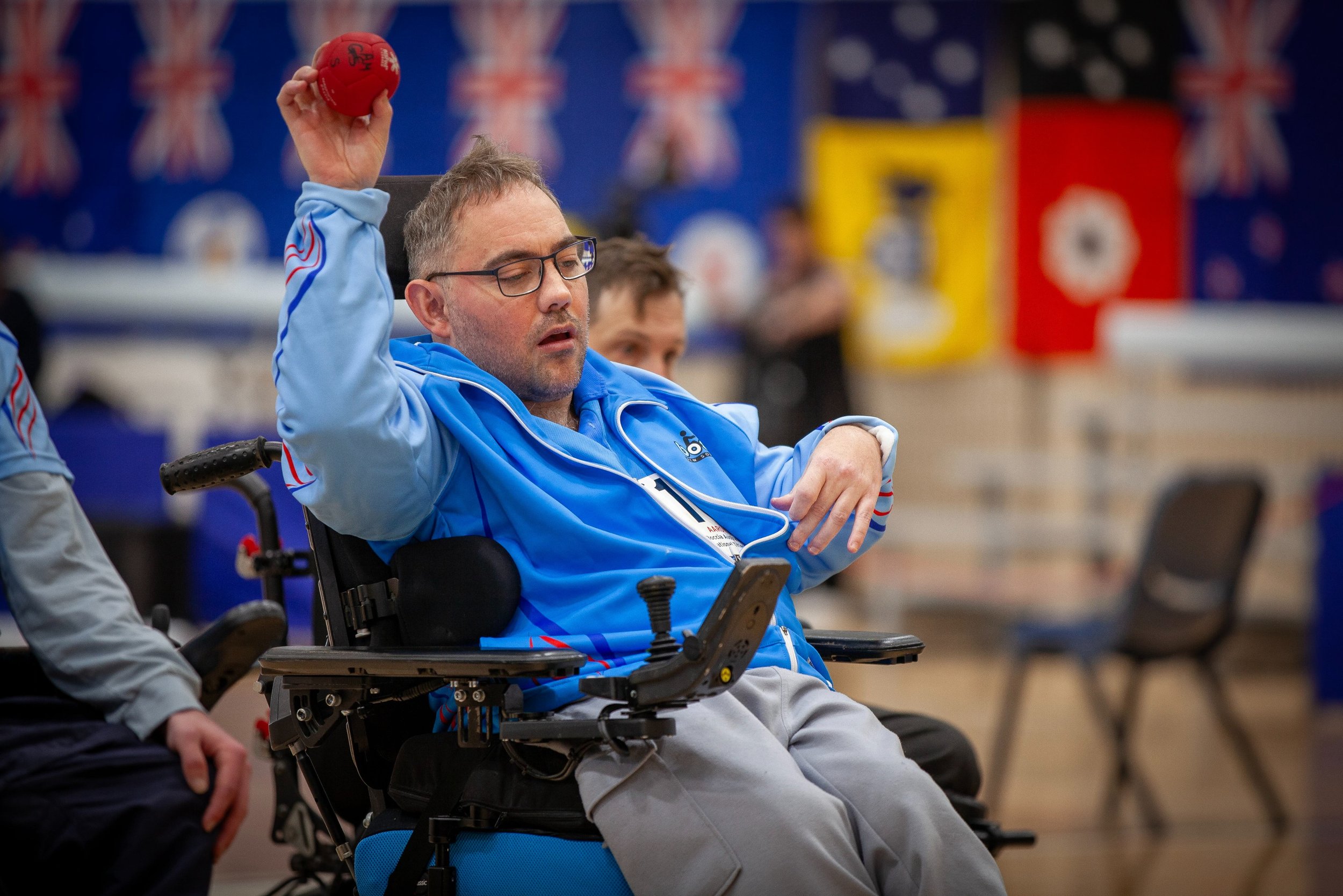 A man in a blue jacket with a disability sports badge, wearing glasses, in a motorized wheelchair holding a red boccia ball in his right hand, during a boccia game in an indoor sports facility.