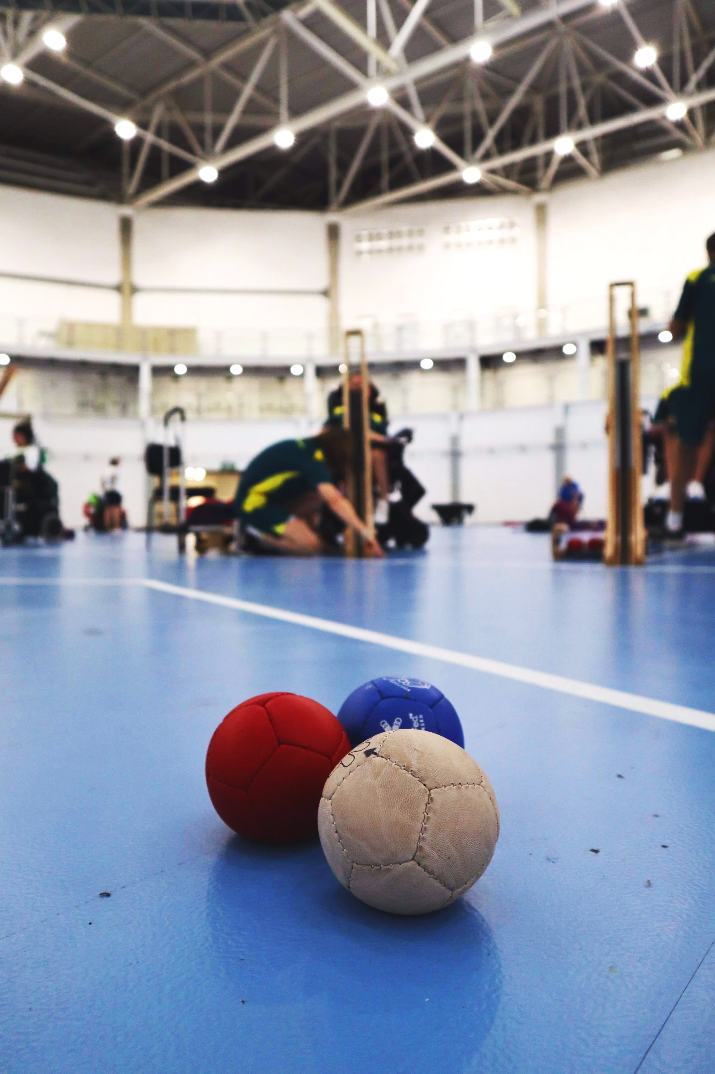 Three boccia—red, blue, and white—rest on a blue indoor boccia court with an indoor gym in the background, featuring players preparing for a game.