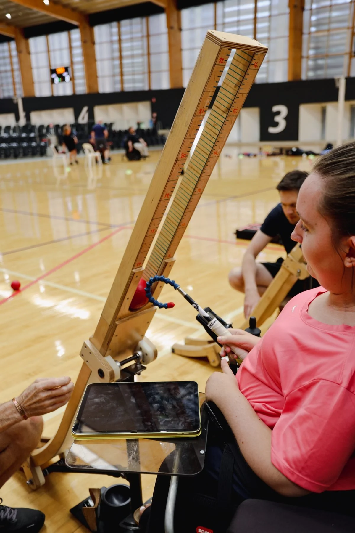 A person in a pink shirt is operating a long, inclined wooden boccia ramp, inside a gymnasium with wooden walls and a boccia court. Several people are sitting and standing in the background.