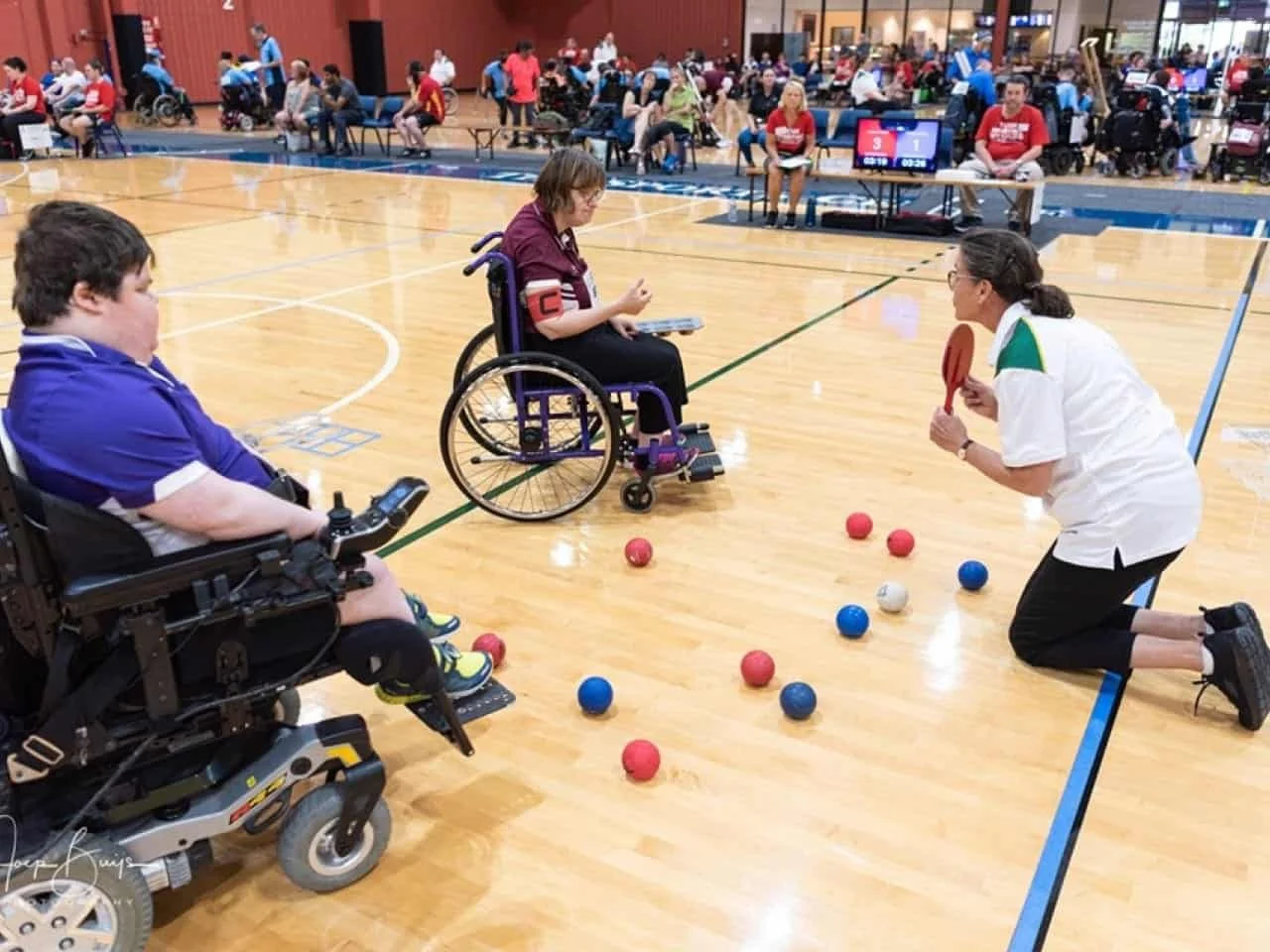 A referee kneeling on a boccia court with two boccia players in wheelchairs, surrounded by an audience in a gymnasium.