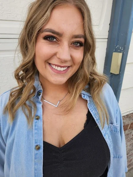 A young woman with wavy blonde hair smiling, wearing a black top, a light blue denim shirt, and a silver necklace, standing outdoors in front of a white wall and a blue door.