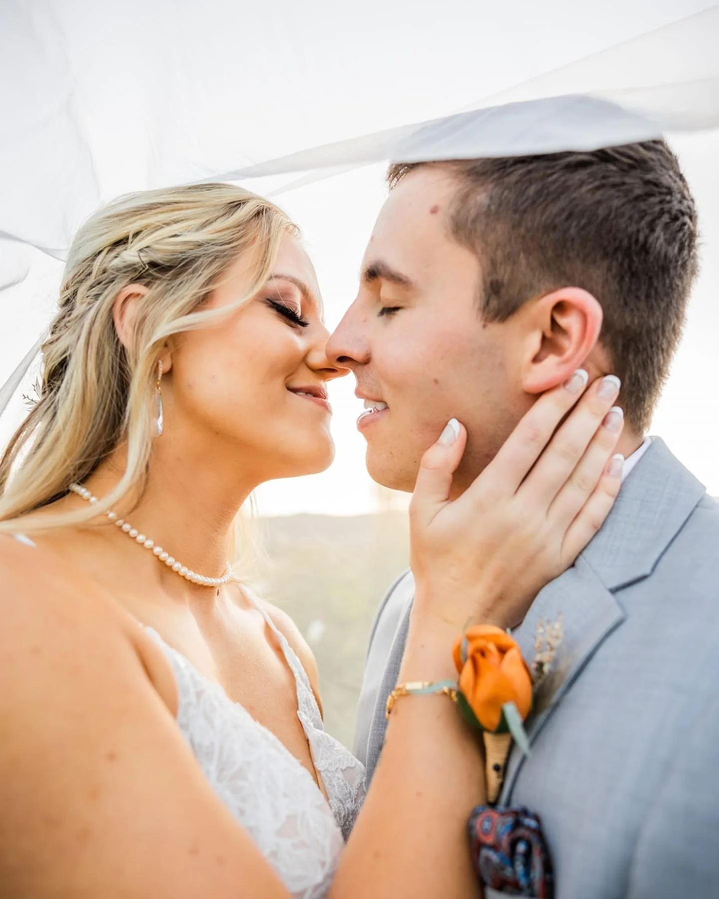 A bride and groom with eyes closed, touching noses and smiling, sharing a close moment outdoors. The bride has blonde hair, pearl necklace, and lace dress, while the groom wears a gray suit with a boutonniere.