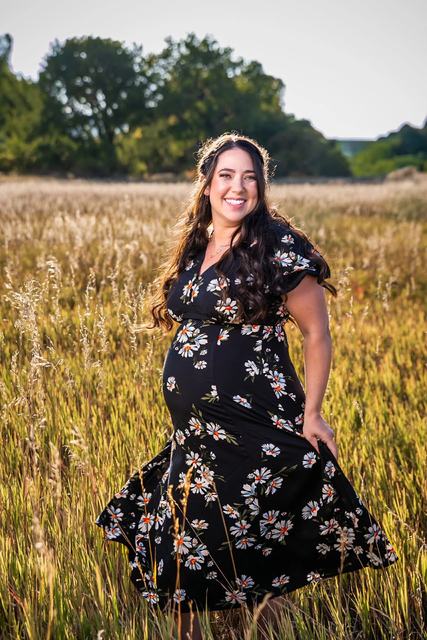A smiling woman with long dark hair, wearing a black floral dress, standing in a grassy field during sunset.