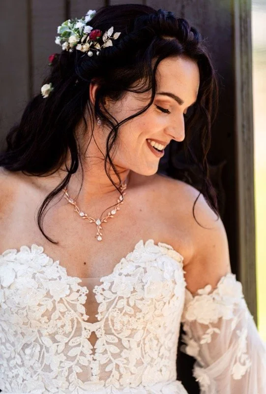 A bride with dark wavy hair decorated with white and red flowers, wearing a white lace wedding dress with off-shoulder sleeves and a pearl necklace, smiling and looking down.