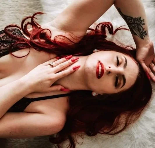 Woman with red hair and red lipstick lying on her back on a soft surface, eyes closed, hand on her neck, other hand resting on her head, tattoo on her arm.