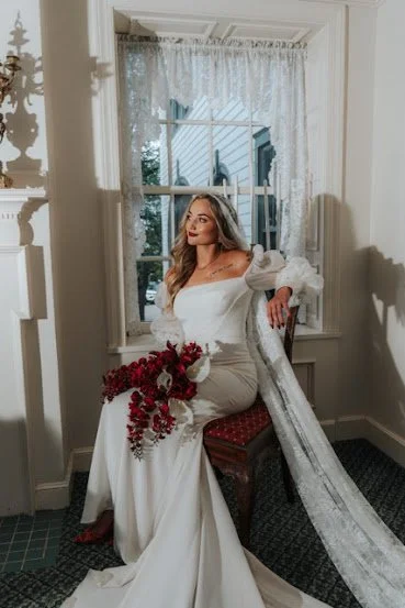 A woman in a white dress sitting on a chair, holding a bouquet of red flowers, in front of a window with lace curtains.