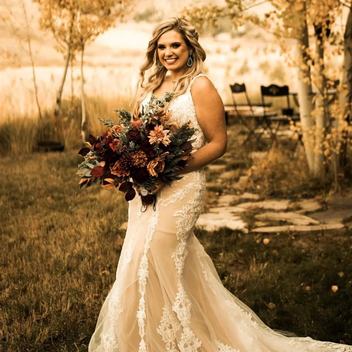 A smiling woman in a white lace wedding dress holding a large bouquet of flowers outdoors during autumn.