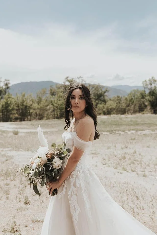 A woman in a white wedding dress holding a bouquet of flowers outdoors, with a mountainous landscape and cloudy sky in the background.
