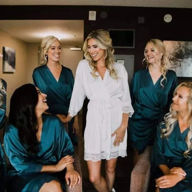 Bridal party with the bride in a white dress and bridesmaids in teal robes, smiling and interacting indoors.