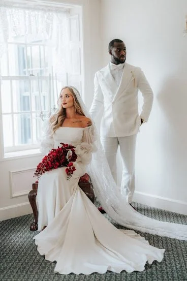 A bride sitting on a bench holding a bouquet, and a groom standing beside her in a white suit, in a bright room with large windows.