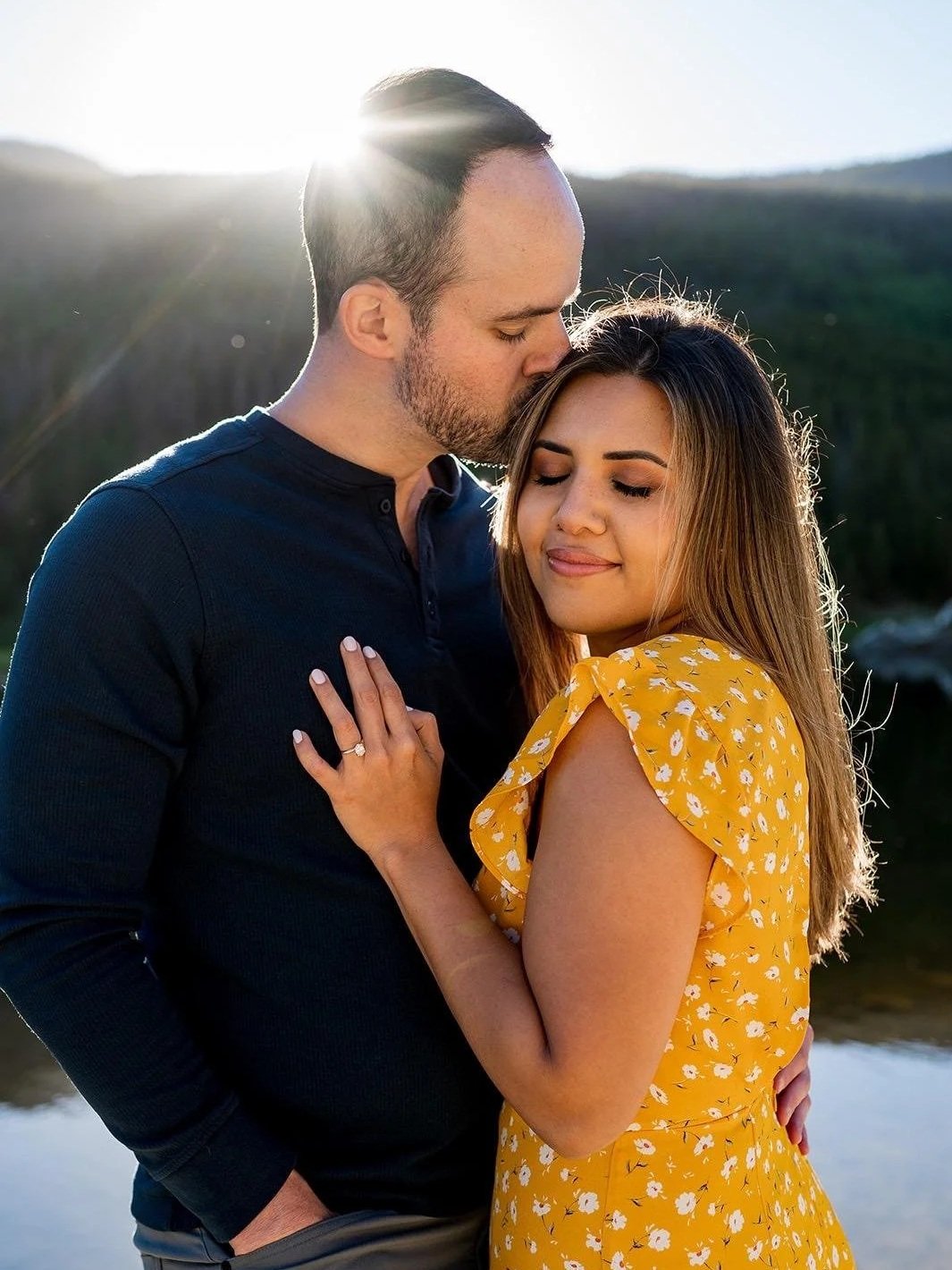 A man kisses a woman on the forehead outdoors with sunlight behind them, near a body of water and green hills.