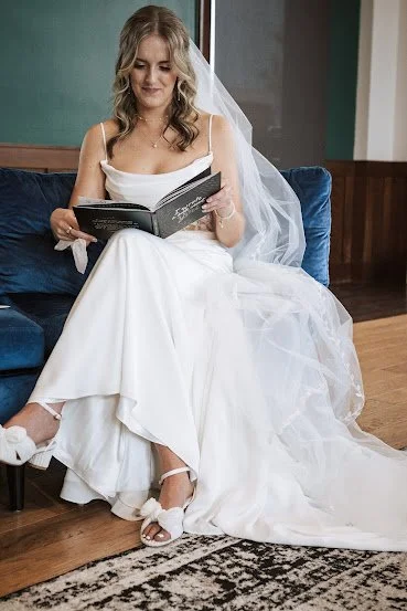 A bride in a white wedding dress with a veil, sitting on a blue couch and reading a book.