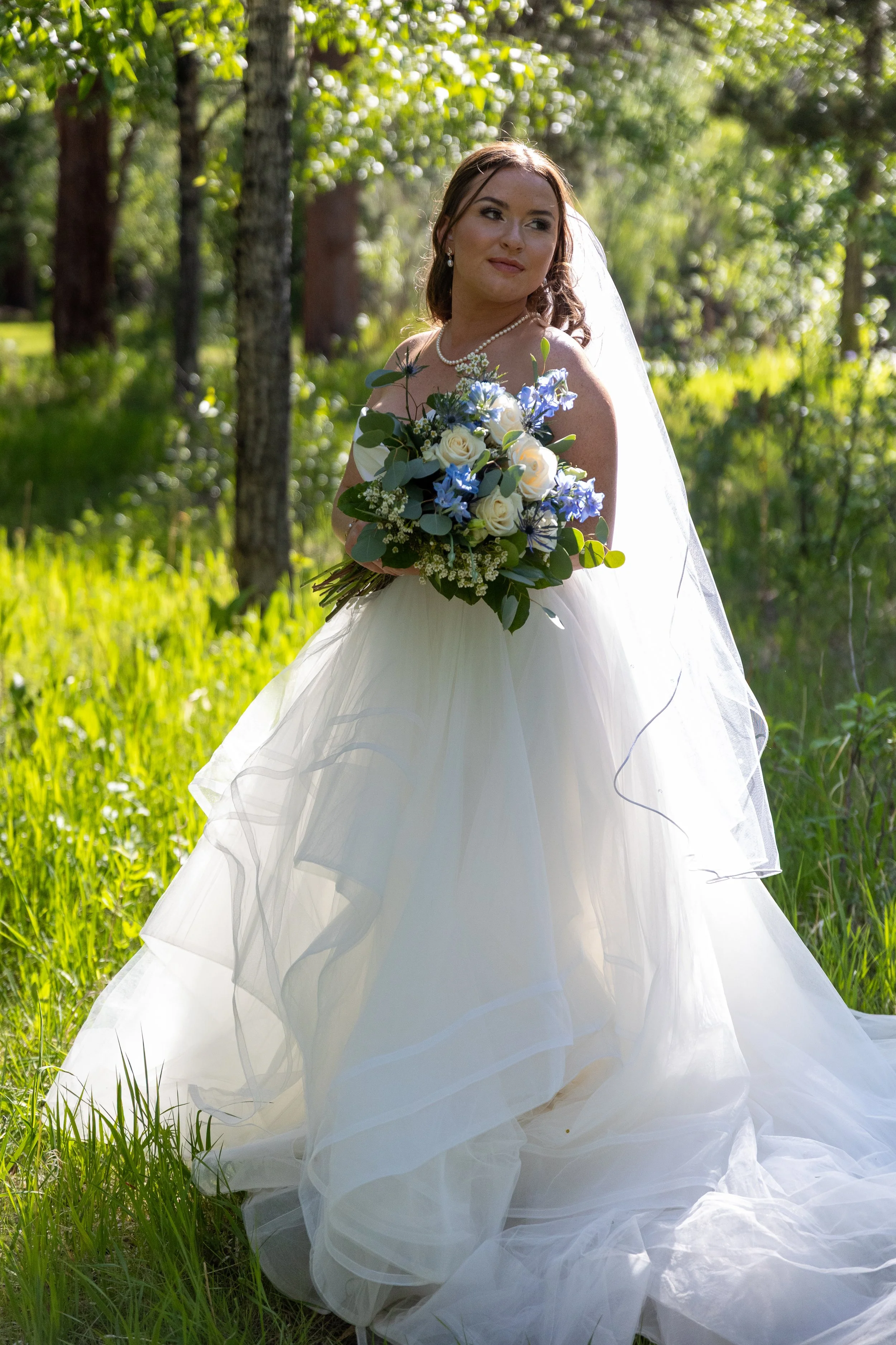 A bride in a white wedding gown holding a bouquet of white and blue flowers, standing in a lush green forest.
