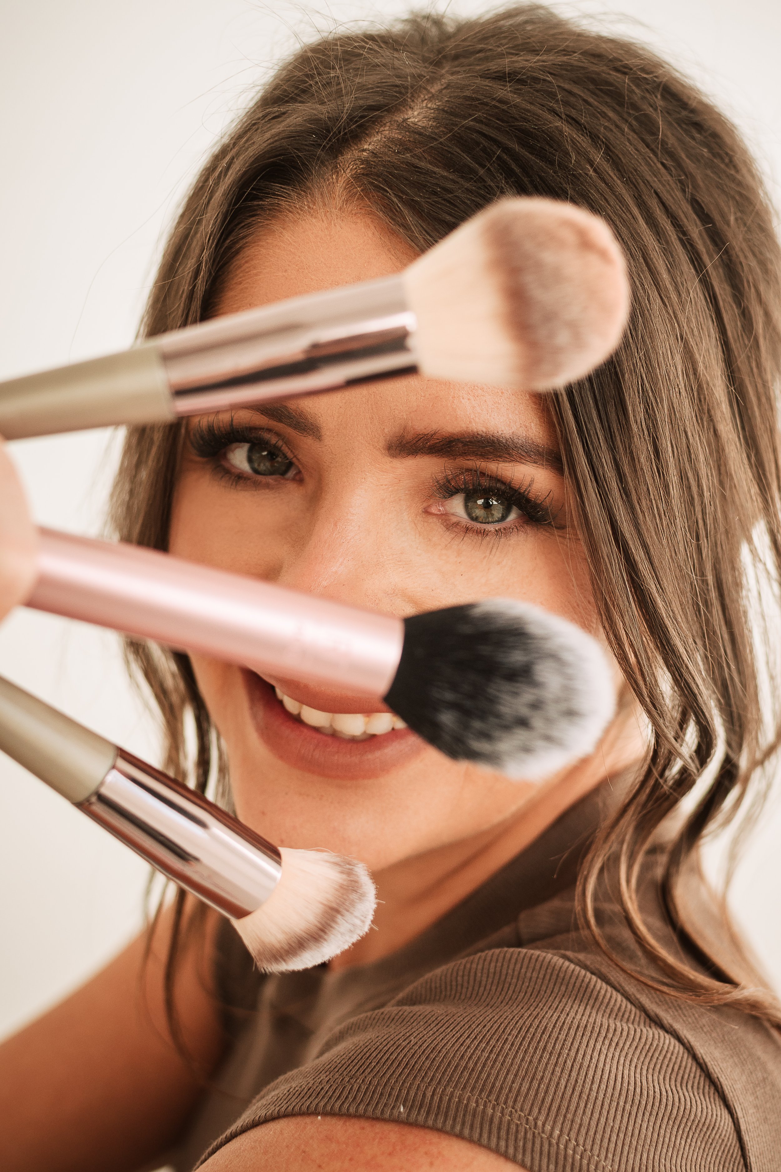 A woman with brown hair and makeup, smiling, holding various makeup brushes in front of her face.