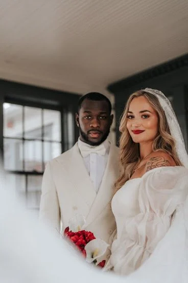 Bride and groom standing indoors in wedding attire, with windows in the background.