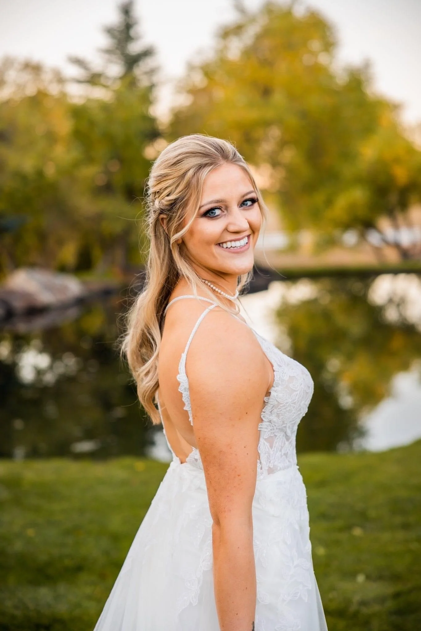 A bride in a white lace wedding dress, smiling outdoors near a pond with trees in the background during sunset.