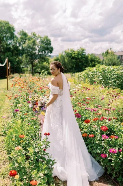 A woman in a white wedding dress holding a bouquet in a colorful flower field outdoors.