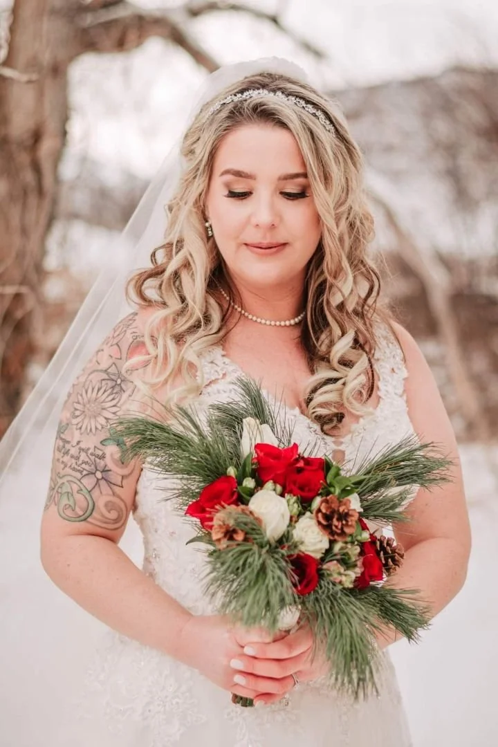 Bride in a white wedding dress with blonde curly hair holding a bouquet of red, white, and pinecone decorations, standing outdoors with blurred trees in the background.