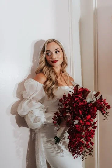 Woman in white dress holding a red floral bouquet, standing against a white wall