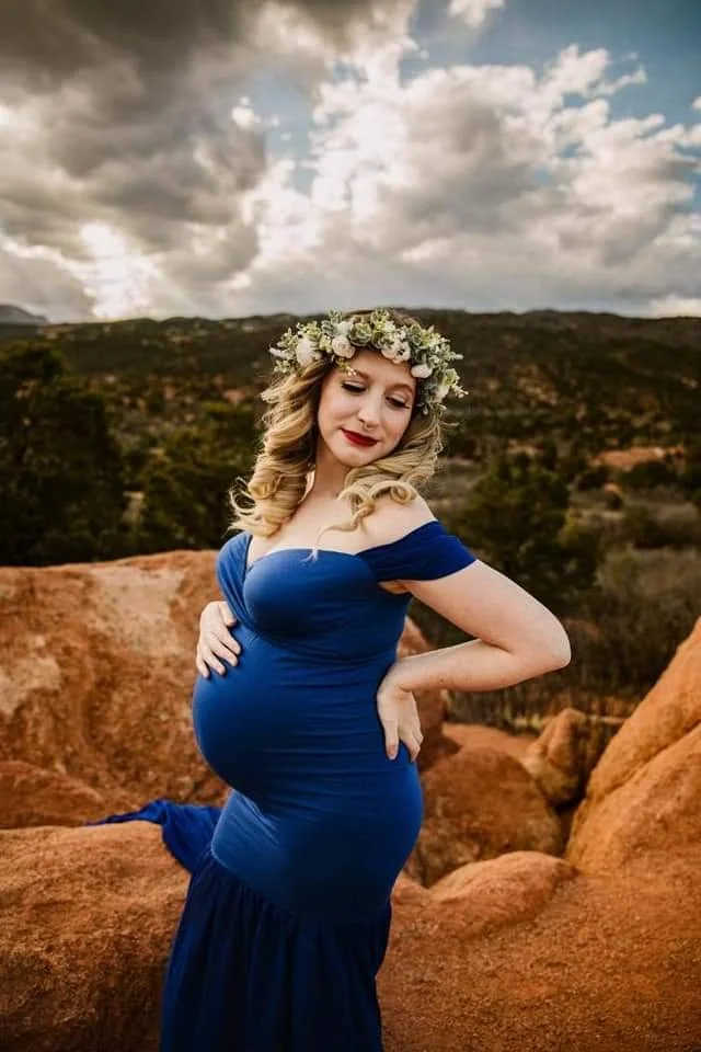 Pregnant woman in a blue off-shoulder dress with a floral crown, standing outdoors on rocky terrain with a cloudy sky in the background.