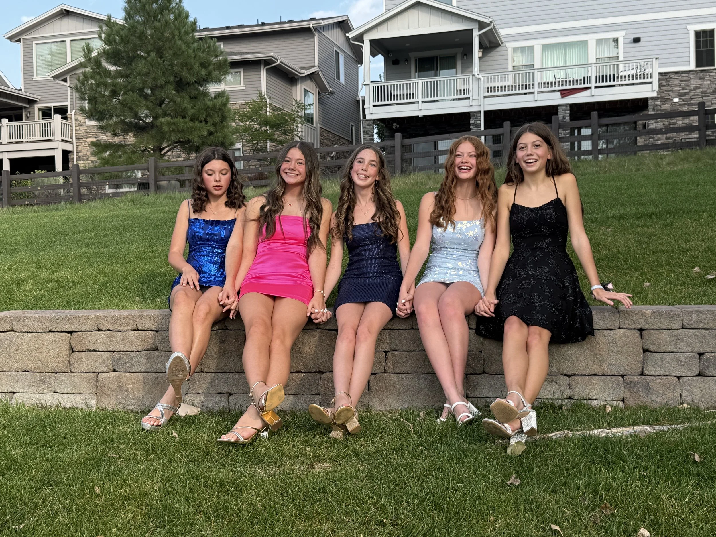 Five young women in colorful dresses sitting on a stone wall, holding hands, with houses and trees in the background.