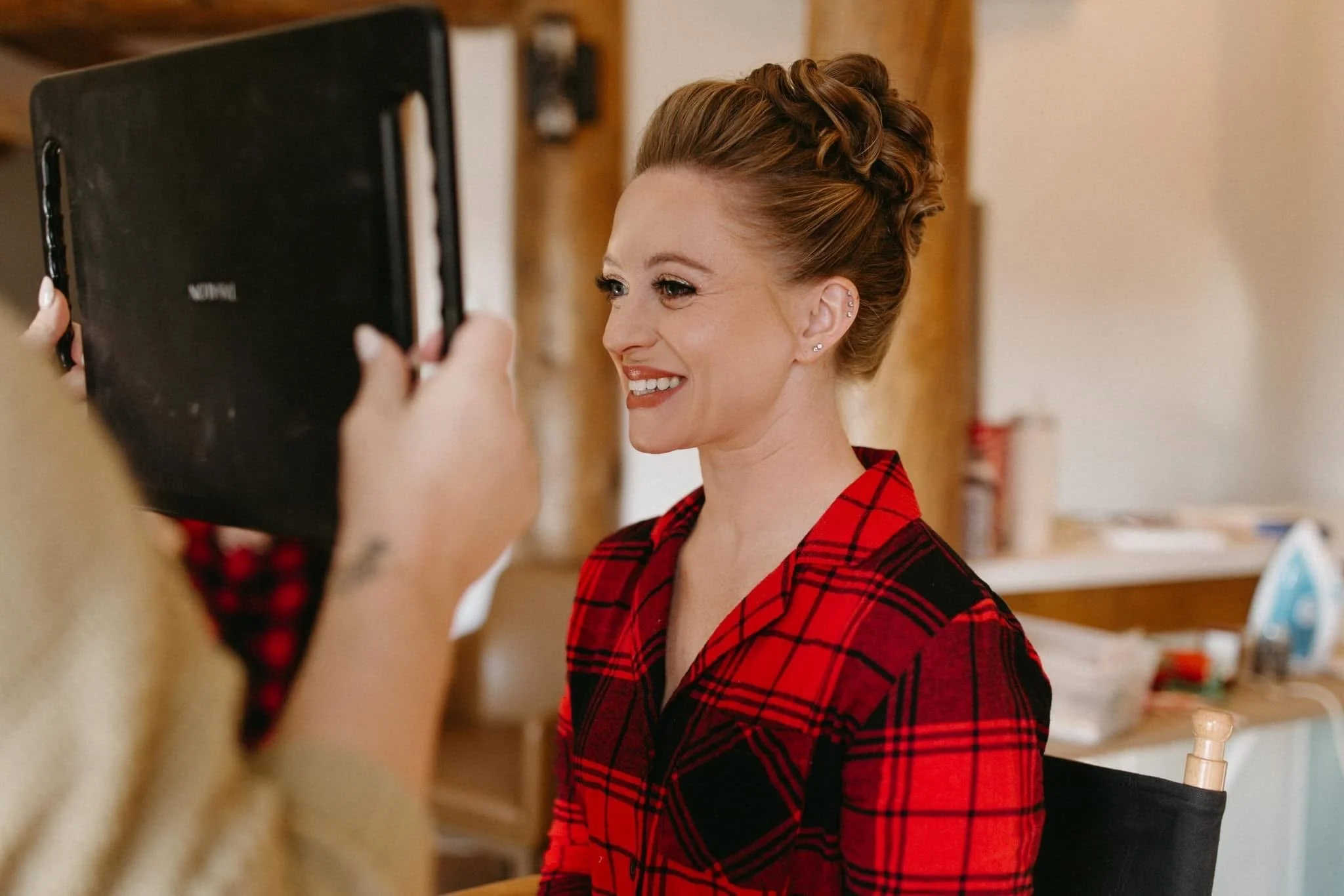A woman in a red plaid shirt is smiling while having her portrait taken indoors.