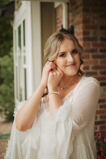 A young woman with blonde hair in a loose braid, wearing a white lace dress and jewelry, smiling and posing outside a brick building.